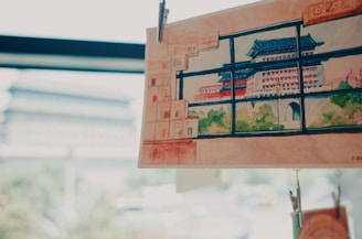 A detailed watercolor painting of a traditional Asian pagoda with green and red accents is clipped and displayed against a softly blurred background. The scene outside the window is reminiscent of the depicted architecture, suggesting a connection between the artwork and its surroundings.