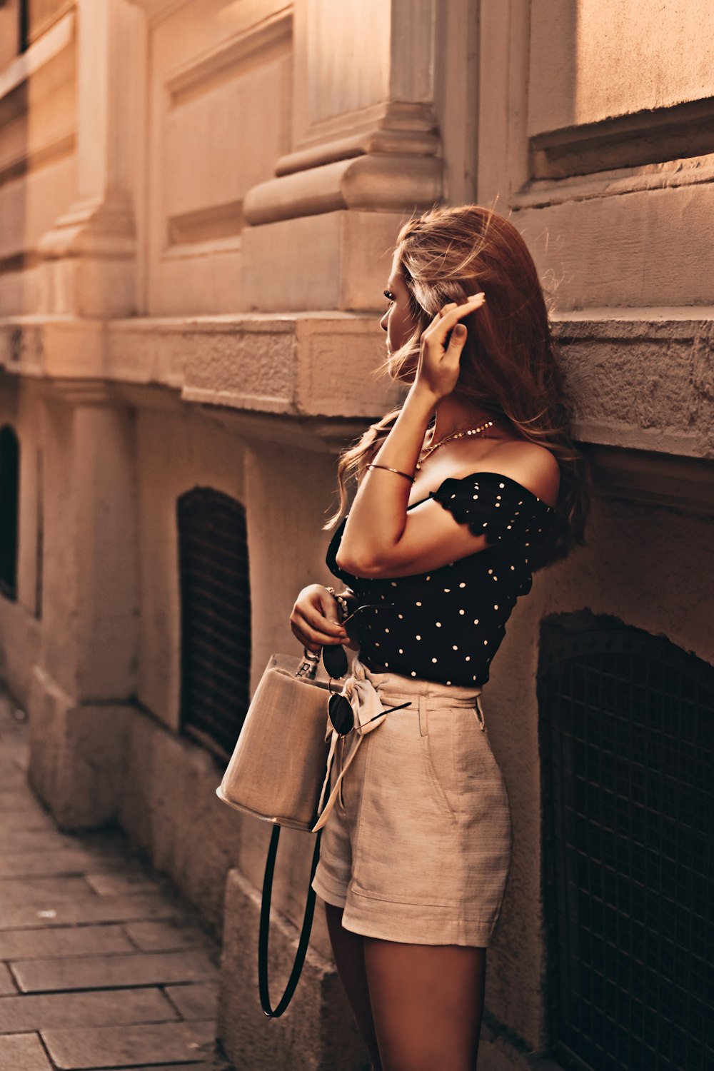 woman wearing black blouse and white short-shorts carrying bag leaning on wall