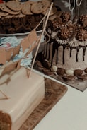 A variety of desserts are displayed on a table. A cake with chocolate drip and Ferrero Rocher toppings is accompanied by a rectangular cake with a fondant finish and decorative flags. In the background, cookies and brownies are arranged on a platter.