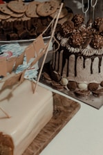 A variety of desserts are displayed on a table. A cake with chocolate drip and Ferrero Rocher toppings is accompanied by a rectangular cake with a fondant finish and decorative flags. In the background, cookies and brownies are arranged on a platter.