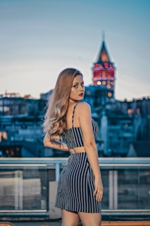 A stylish woman in a flowing dress standing on an urban rooftop at sunset, the city skyline blurred in the background