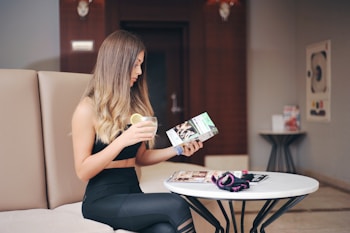 A woman with long hair is sitting on a beige couch in a modern indoor setting. She is holding a drink with a lemon slice in one hand and is reading a magazine or brochure with the other. She is wearing athletic clothing, and on the table in front of her are a few magazines and a small item of clothing, possibly a sports accessory. The room features neutral tones with dark wood accents.