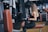A woman is seated on a workout bench using a gym machine. She is wearing black athletic wear and gloves, and appears focused on her exercise in a modern gym environment. Various gym equipment is visible in the background.