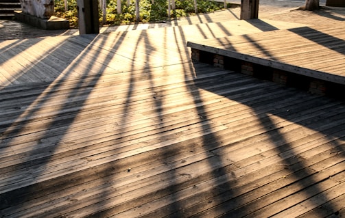 Wooden decking is bathed in warm sunlight, casting long, defined shadows from vertical beams. The light creates a pattern on the textured surface, adding depth and dimension to the scene.