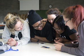 A group of young people are gathered around a table, looking intently at a smartphone. They appear engaged and are smiling, indicating enjoyment or interest in what they are viewing. The setting is likely a classroom or similar environment, suggested by the presence of bags and a clock on the wall in the background. Casual clothing suggests a relaxed atmosphere.