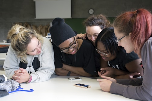A group of young people are gathered around a table, looking intently at a smartphone. They appear engaged and are smiling, indicating enjoyment or interest in what they are viewing. The setting is likely a classroom or similar environment, suggested by the presence of bags and a clock on the wall in the background. Casual clothing suggests a relaxed atmosphere.