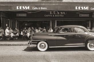 A vintage car is parked in front of a cafe with people sitting at outdoor tables, enjoying a meal or drinks. The setting has a classic and nostalgic feel.