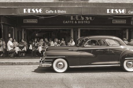 A vintage car is parked in front of a cafe with people sitting at outdoor tables, enjoying a meal or drinks. The setting has a classic and nostalgic feel.