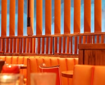 The image depicts an interior of a restaurant or dining area with orange cushioned seats and wooden tables. There are vertical wooden slats which serve as partitions, behind which a blue wall or window is visible. The overall setting has a cozy and warm ambiance.