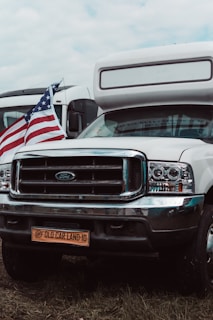 A white camper van with a Ford emblem is parked on grass, displaying an American flag on the front. The license plate reads 'Old Car Land-10'. Another vehicle is visible in the background.