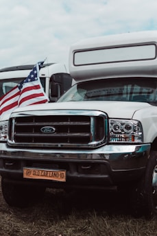 A white camper van with a Ford emblem is parked on grass, displaying an American flag on the front. The license plate reads 'Old Car Land-10'. Another vehicle is visible in the background.