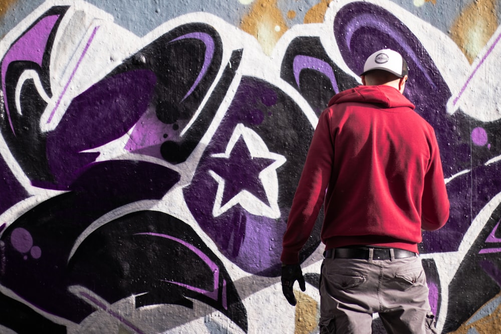 man wearing red pullover hoodie and denim jeans standing beside graffiti