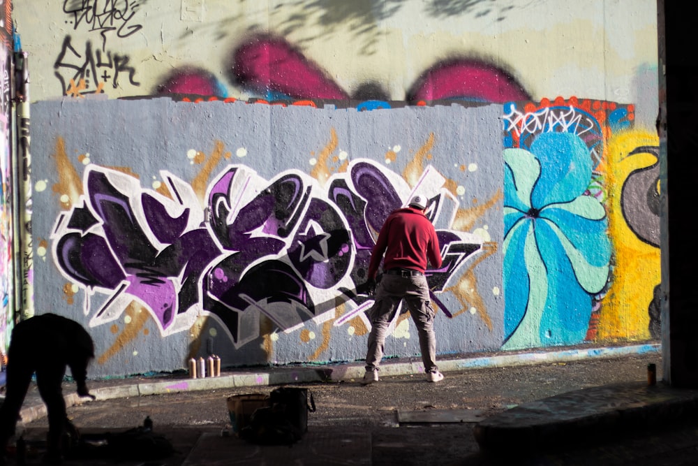 man wearing red jacket standing while painting on concrete wall during daytime