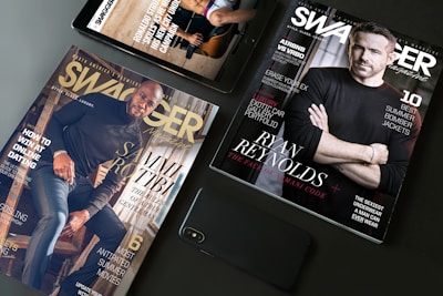 A flat lay arrangement featuring two Swagger magazines and a smartphone on a table. One magazine displays an image of a man in a black shirt, while the other features another man sitting on stairs. Both magazines have text highlighting fashion tips and lifestyle content.