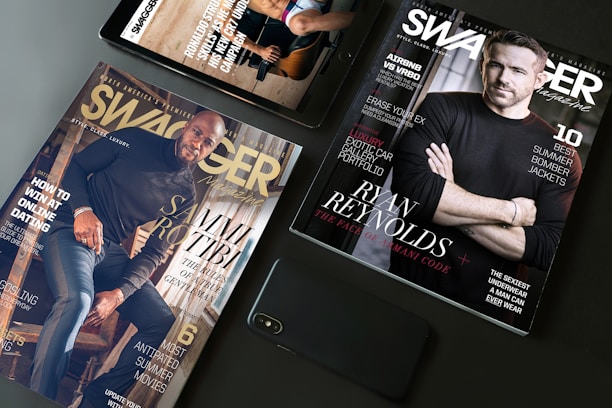 A flat lay arrangement featuring two Swagger magazines and a smartphone on a table. One magazine displays an image of a man in a black shirt, while the other features another man sitting on stairs. Both magazines have text highlighting fashion tips and lifestyle content.