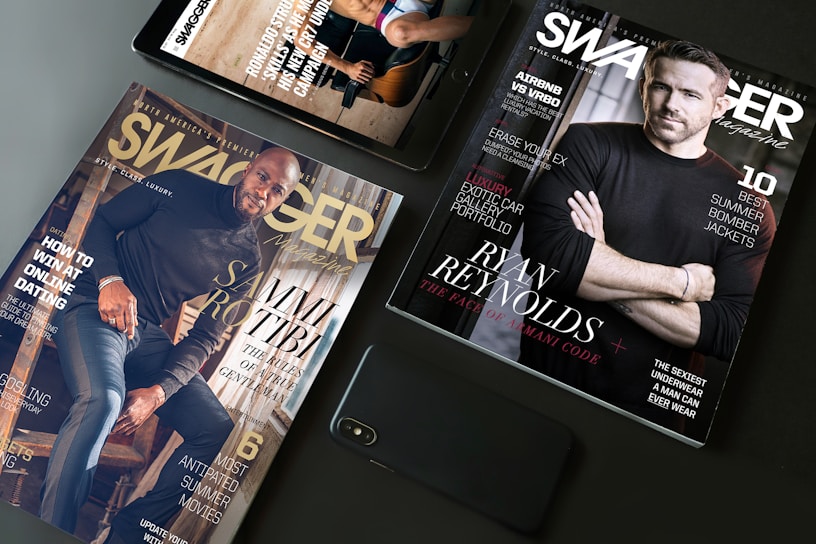 A flat lay arrangement featuring two Swagger magazines and a smartphone on a table. One magazine displays an image of a man in a black shirt, while the other features another man sitting on stairs. Both magazines have text highlighting fashion tips and lifestyle content.