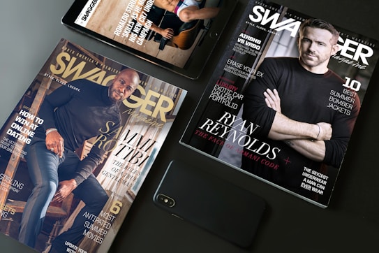 A flat lay arrangement featuring two Swagger magazines and a smartphone on a table. One magazine displays an image of a man in a black shirt, while the other features another man sitting on stairs. Both magazines have text highlighting fashion tips and lifestyle content.