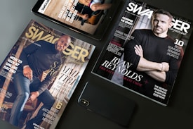A flat lay arrangement featuring two Swagger magazines and a smartphone on a table. One magazine displays an image of a man in a black shirt, while the other features another man sitting on stairs. Both magazines have text highlighting fashion tips and lifestyle content.