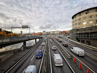 A multi-lane highway with numerous vehicles traveling in both directions. The highway is elevated, with railings separating the lanes. On the right, there is a large, curved building and some construction cones on the road. The sky is overcast with grey clouds, and there are buildings with various architectural styles in the distance. The left side shows a body of water lined with trees with autumn foliage.