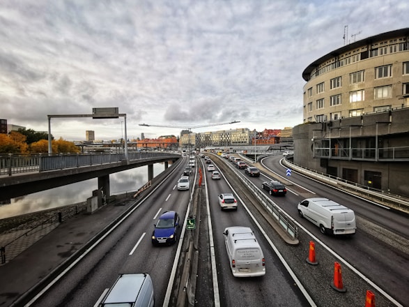 A multi-lane highway with numerous vehicles traveling in both directions. The highway is elevated, with railings separating the lanes. On the right, there is a large, curved building and some construction cones on the road. The sky is overcast with grey clouds, and there are buildings with various architectural styles in the distance. The left side shows a body of water lined with trees with autumn foliage.