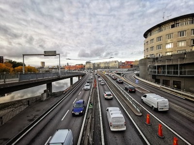 A multi-lane highway with numerous vehicles traveling in both directions. The highway is elevated, with railings separating the lanes. On the right, there is a large, curved building and some construction cones on the road. The sky is overcast with grey clouds, and there are buildings with various architectural styles in the distance. The left side shows a body of water lined with trees with autumn foliage.