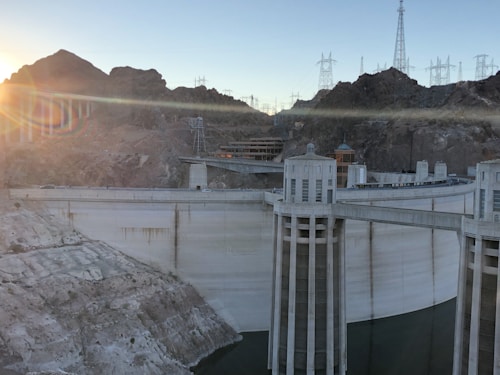 A large, concrete dam with cylindrical intake towers and a spillway constructed in a rocky canyon environment. High voltage power lines and towers are visible in the background, with sunlight streaming over the mountain tops.