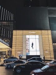 A bustling urban street scene at night, featuring a large illuminated advertisement for Cartier on the side of a building. Several cars are visible on the road, and a group of people stand near the entrance, which reads Bukit Bintang Entrance. The overall ambiance is lively and modern, with glowing lights and tall buildings in the background.