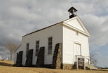 An old white wooden church with a steeple and a cross on top, located in a rural area with a cloudy sky in the background. The church is supported by stone pillars and has a simple sign indicating 'Saint Catherine's Catholic Church' along with service times. The surrounding environment includes a barren tree and some distant tombstones, suggesting a nearby cemetery.