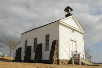 An old white wooden church with a steeple and a cross on top, located in a rural area with a cloudy sky in the background. The church is supported by stone pillars and has a simple sign indicating 'Saint Catherine's Catholic Church' along with service times. The surrounding environment includes a barren tree and some distant tombstones, suggesting a nearby cemetery.
