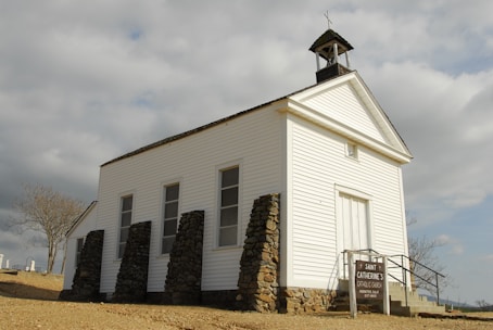 An old white wooden church with a steeple and a cross on top, located in a rural area with a cloudy sky in the background. The church is supported by stone pillars and has a simple sign indicating 'Saint Catherine's Catholic Church' along with service times. The surrounding environment includes a barren tree and some distant tombstones, suggesting a nearby cemetery.