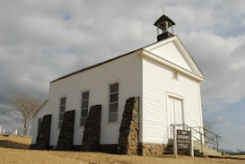 An old white wooden church with a steeple and a cross on top, located in a rural area with a cloudy sky in the background. The church is supported by stone pillars and has a simple sign indicating 'Saint Catherine's Catholic Church' along with service times. The surrounding environment includes a barren tree and some distant tombstones, suggesting a nearby cemetery.