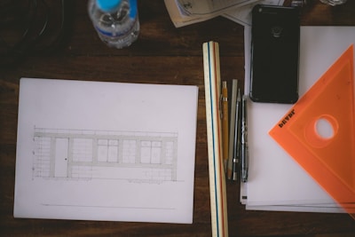 A wooden table with various drafting and design tools including a sketch of a building, a water bottle, a smartphone, rulers, pens, and an orange drafting triangle. The sketch depicts a facade with windows and a door, while papers and a translucent orange triangle are neatly arranged nearby.