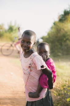 Two children stand on a dirt path, with one child wearing a pink shirt and smiling while the other is carried in a sling, also wearing a pink shirt. The background features blurred greenery and a person riding a bicycle.