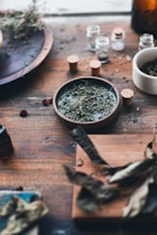 A rustic wooden table adorned with various dried herbs in small corked glass jars and bowls. The setting includes a mix of leaves and herbs scattered around, suggesting an apothecary or herbal preparation scene.