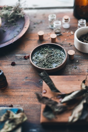 A rustic wooden table adorned with various dried herbs in small corked glass jars and bowls. The setting includes a mix of leaves and herbs scattered around, suggesting an apothecary or herbal preparation scene.