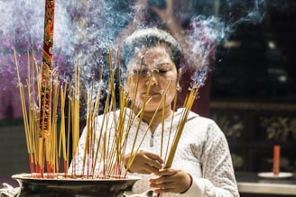 A woman is holding and arranging incense sticks in a large, ornate vessel. Smoke softly drifts upward from the burning incense, creating a serene and spiritual atmosphere. The woman's expression appears focused and calm, with the background slightly blurred to emphasize her contemplative state.