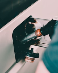 A close-up view of a fuel pump nozzle inserted into a vehicle's fuel tank, with a cap resting nearby. The image shows rust and metal around the opening.