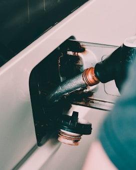 A close-up view of a fuel pump nozzle inserted into a vehicle's fuel tank, with a cap resting nearby. The image shows rust and metal around the opening.