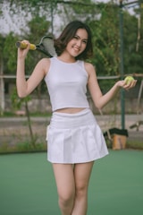 A young woman stands on a tennis court, smiling confidently. She holds a tennis racket over one shoulder and a tennis ball in her other hand. She is wearing a white tank top and a white tennis skirt.