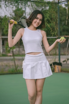 A young woman stands on a tennis court, smiling confidently. She holds a tennis racket over one shoulder and a tennis ball in her other hand. She is wearing a white tank top and a white tennis skirt.