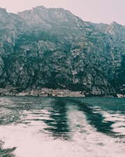 A vast and imposing mountain range rises steeply from the shoreline, with rocky and rugged textures. Below, the calm sea reflects the mountains, creating a mirrored effect above the white frothy waves generated by a moving boat.