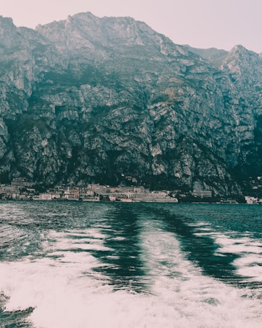 A vast and imposing mountain range rises steeply from the shoreline, with rocky and rugged textures. Below, the calm sea reflects the mountains, creating a mirrored effect above the white frothy waves generated by a moving boat.