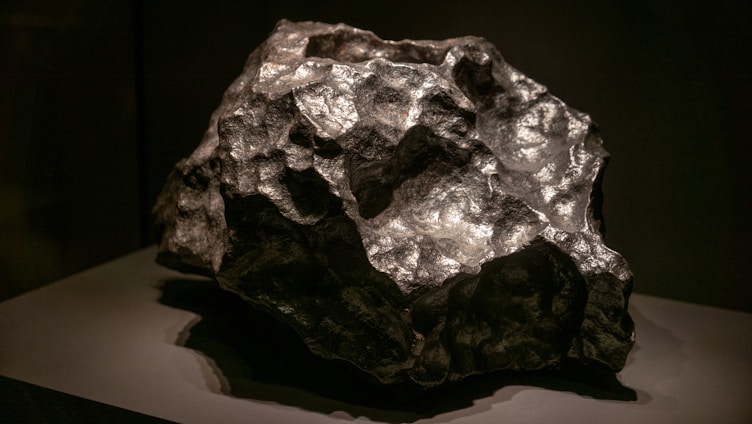 A large, rough-textured meteorite with a metallic, reflective surface. It has an irregular shape and is displayed on a dark, flat surface, highlighting its natural contours and mineral composition.