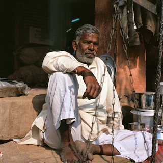A candid photo of an elderly man standing barefoot on a sunlit wooden floor, embodying raw simplicity.