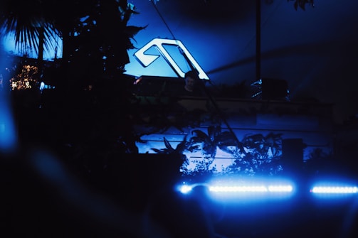 A nighttime scene featuring a person at a DJ booth surrounded by lush greenery. A prominent logo glows in blue on a backdrop behind the DJ. Blue neon lights illuminate the area, creating a moody ambiance.
