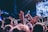 A large crowd of people with their hands raised, possibly enjoying a live music concert. Bright stage lights illuminate the scene, and the focus is on the enthusiastic audience. Some concert-goers wear wristbands, suggesting a festival or a ticketed event.