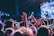 A large crowd of people with their hands raised, possibly enjoying a live music concert. Bright stage lights illuminate the scene, and the focus is on the enthusiastic audience. Some concert-goers wear wristbands, suggesting a festival or a ticketed event.