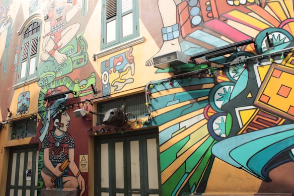 A vibrant mural covering the side of a building features colorful and abstract figures and shapes. The artwork includes a large seated figure with detailed patterns, surrounded by imaginative elements like geometric shapes, plants, and animals in a dynamic composition. Large windows and doors are integrated into the mural, with multicolored string lights hanging below the art.