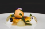 A gourmet dessert with a scoop of orange sorbet placed atop a caramelized custard base. Garnished with fresh mint leaves, edible flowers, and dollops of white cream, accompanied by small melon balls. Served on a white plate with a dark background.