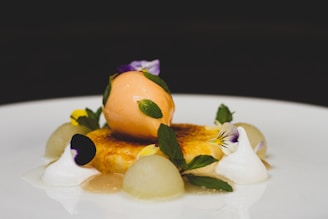 A gourmet dessert with a scoop of orange sorbet placed atop a caramelized custard base. Garnished with fresh mint leaves, edible flowers, and dollops of white cream, accompanied by small melon balls. Served on a white plate with a dark background.